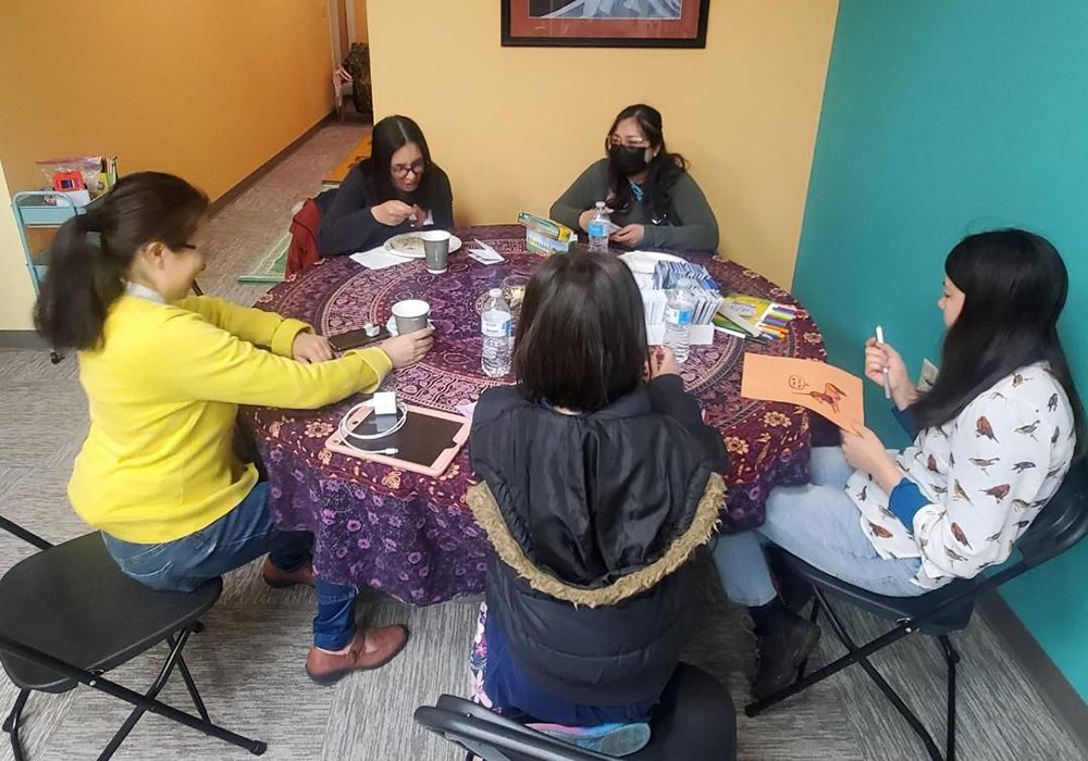 5 people sitting around a round table, all with dark hair, all femmes. one person is wearing a mask.
